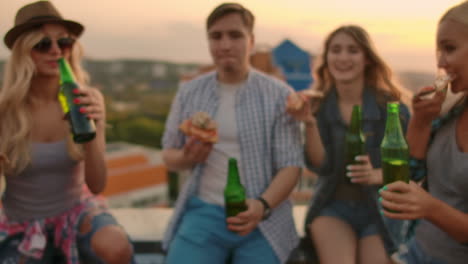 Russian-girls-and-boys-clinks-glasses-and-drinks-beer-from-green-bottels-on-the-party-with-friends-on-the-roof-at-the-sunset.They-are-sitting-together-eat-hot-pizza-after-in-summer-everning.
